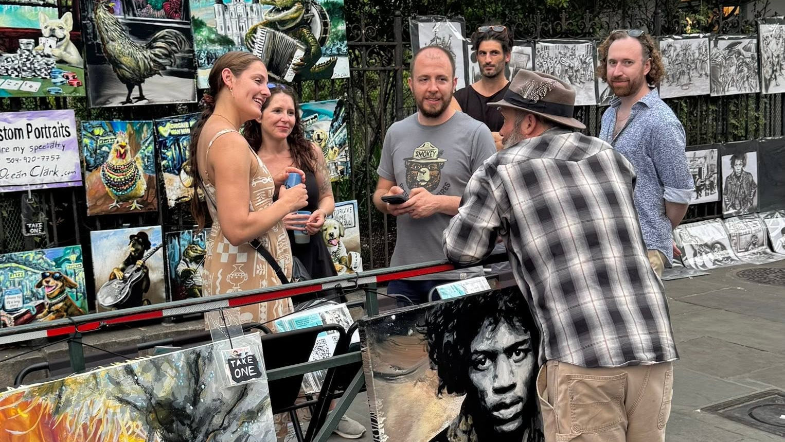 An outdoor street art display with multiple canvases and a large black-and-white portrait painting on a table, surrounded by several people standing and viewing the artwork.