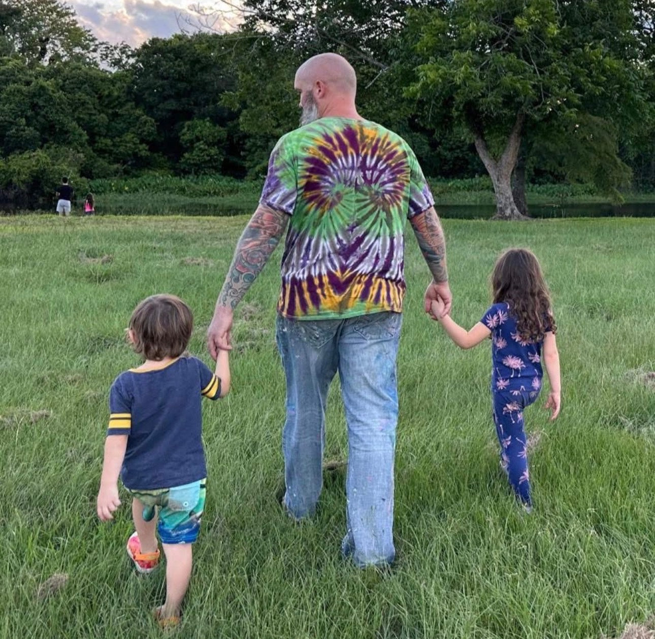 Ocean Clark wearing a colorful spiral tie-dye t-shirt and blue jeans walks hand-in-hand with his two children in a grassy park during the evening.