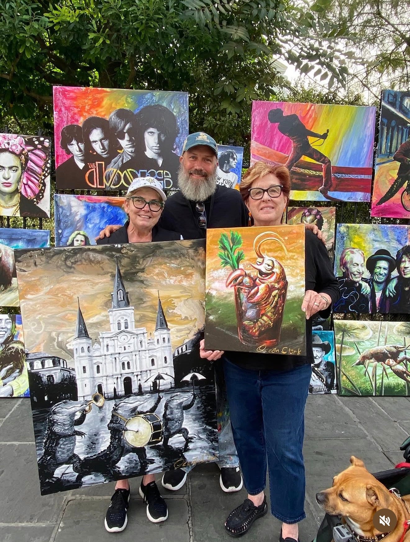 Ocean Clark with two customers standing outdoors in front of a display of colorful paintings, holding up two custom artworks.